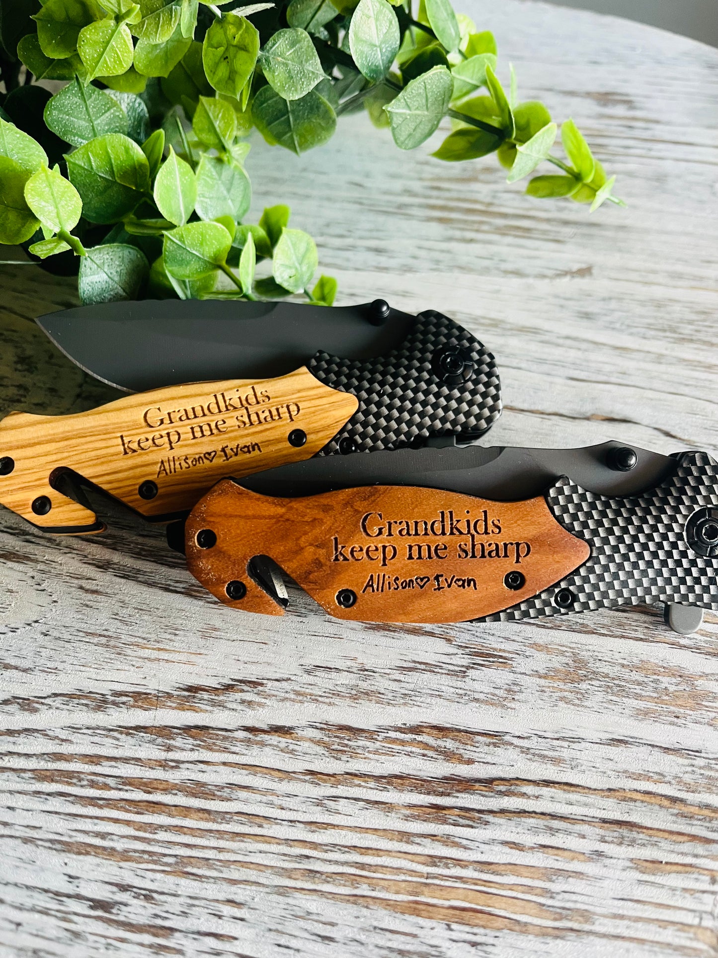 Two folding pocket knives with wooden handles, one with the blade open displaying the engraved text on the handle, the other with the blade closed. Set against a wooden background with green leaf accents.