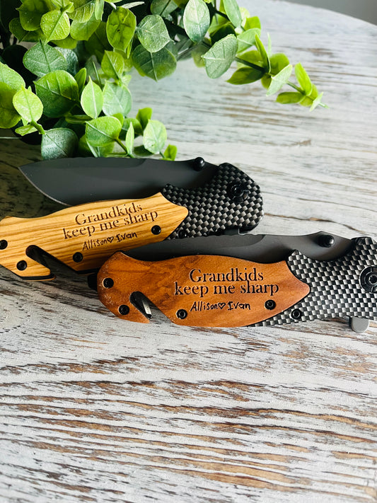 Two folding pocket knives with wooden handles, one with the blade open displaying the engraved text on the handle, the other with the blade closed. Set against a wooden background with green leaf accents.
